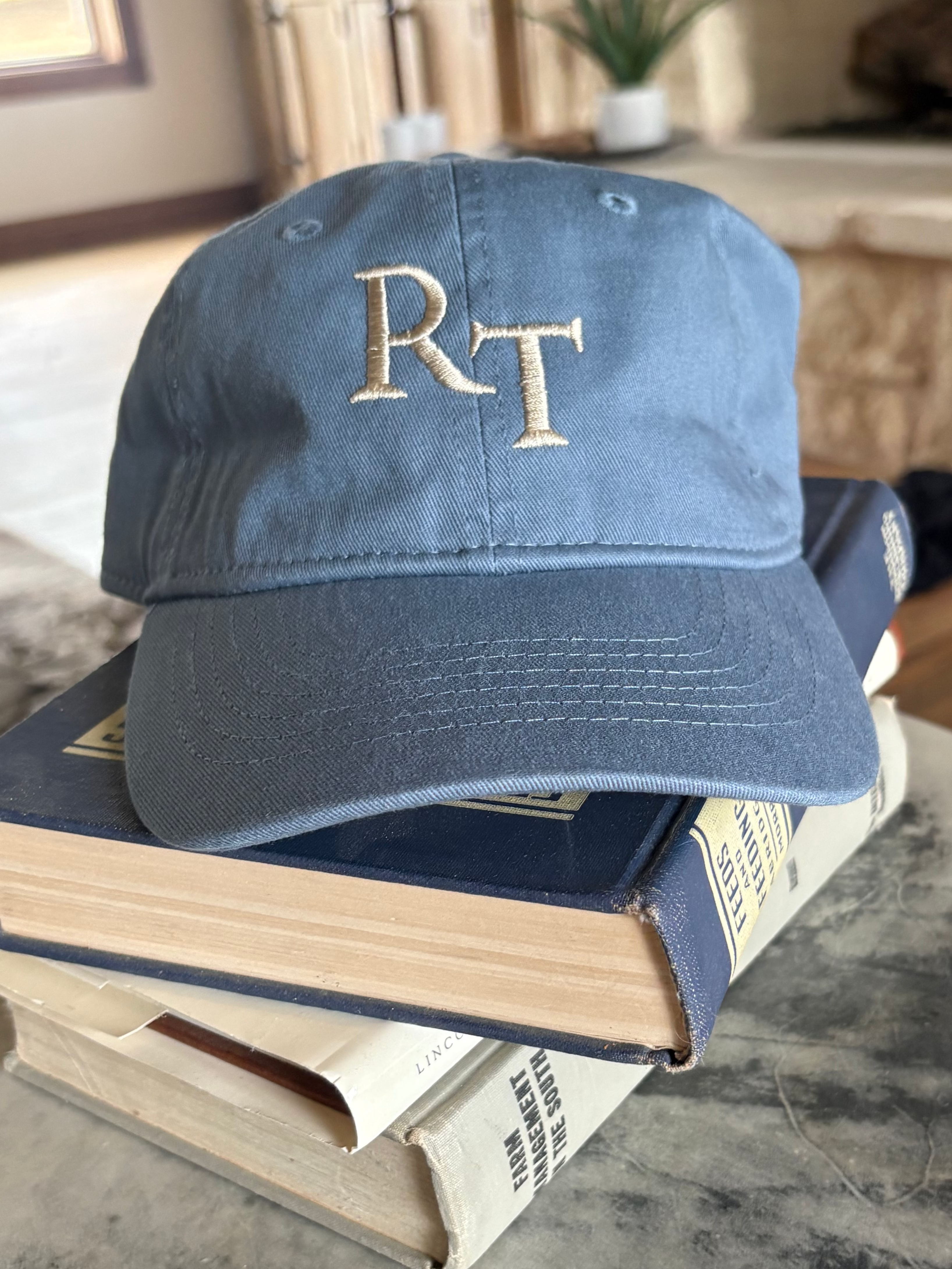 A blue cotton baseball cap with gold embroidered initials 'RT' on the front, resting on a stack of books on a round stone table in a home interior.