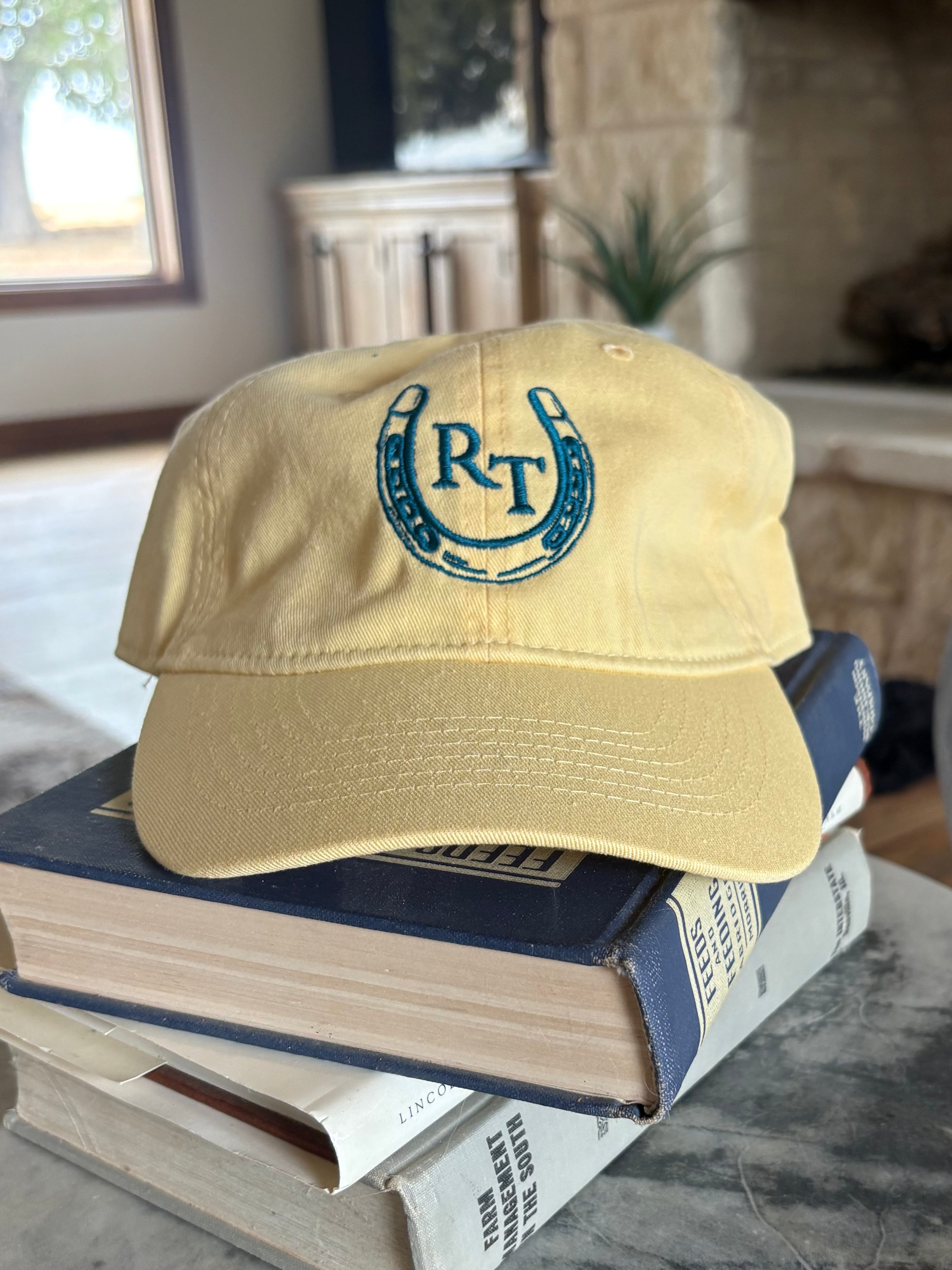 A light yellow baseball cap with a blue embroidered horseshoe logo featuring the letters 'RT' sits on top of a stack of books on a round gray stone table in a home interior.