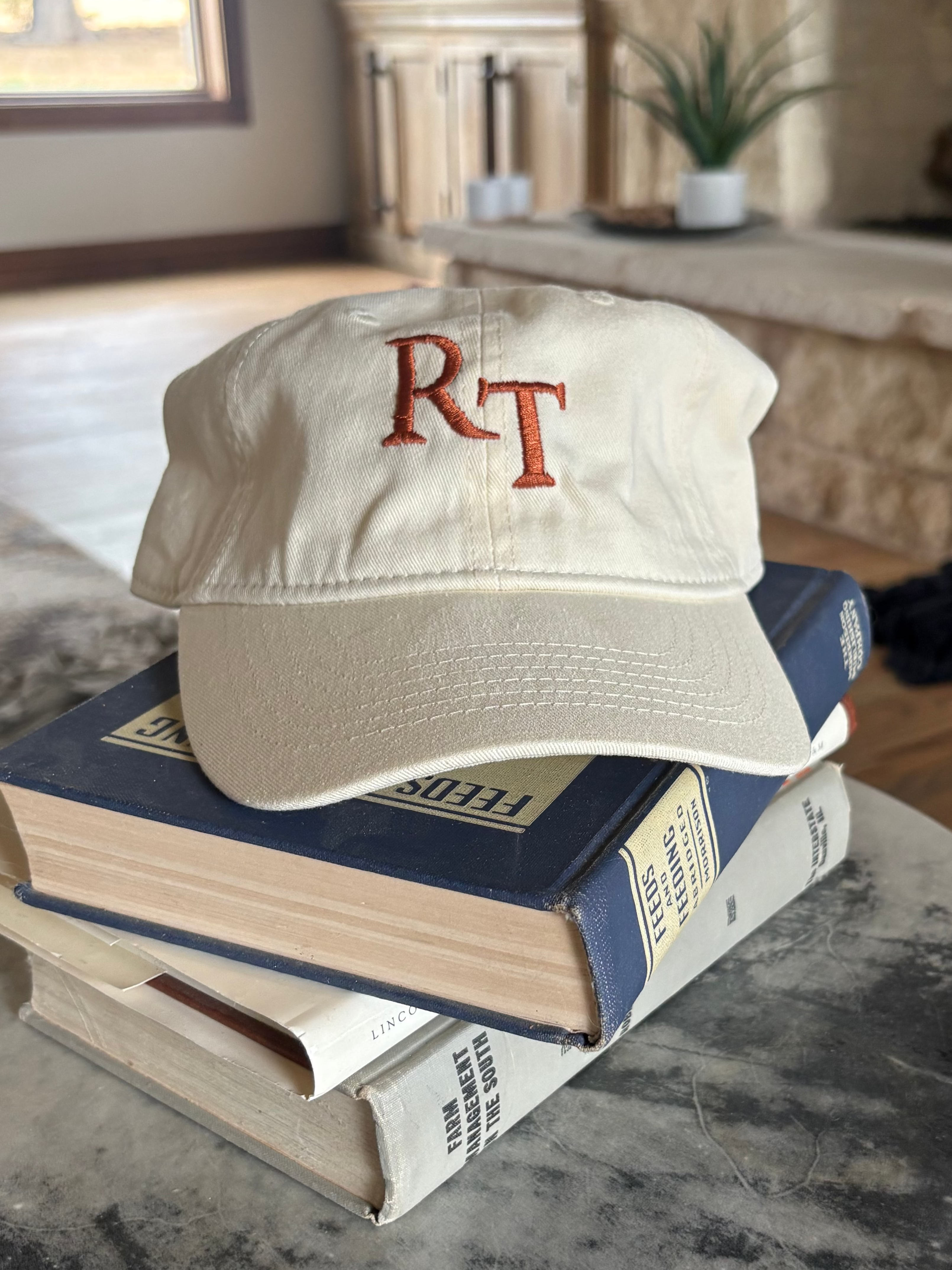 An off-white baseball cap with embroidered red letters 'RT' sits on top of a stack of books on a round gray marble-top table in a living room setting.