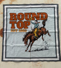 A square bandana featuring a western-themed graphic print with the text 'ROUND TOP EST. 1868' and an illustration of a cowboy riding a bucking brown horse, laid flat on a textured beige surface.