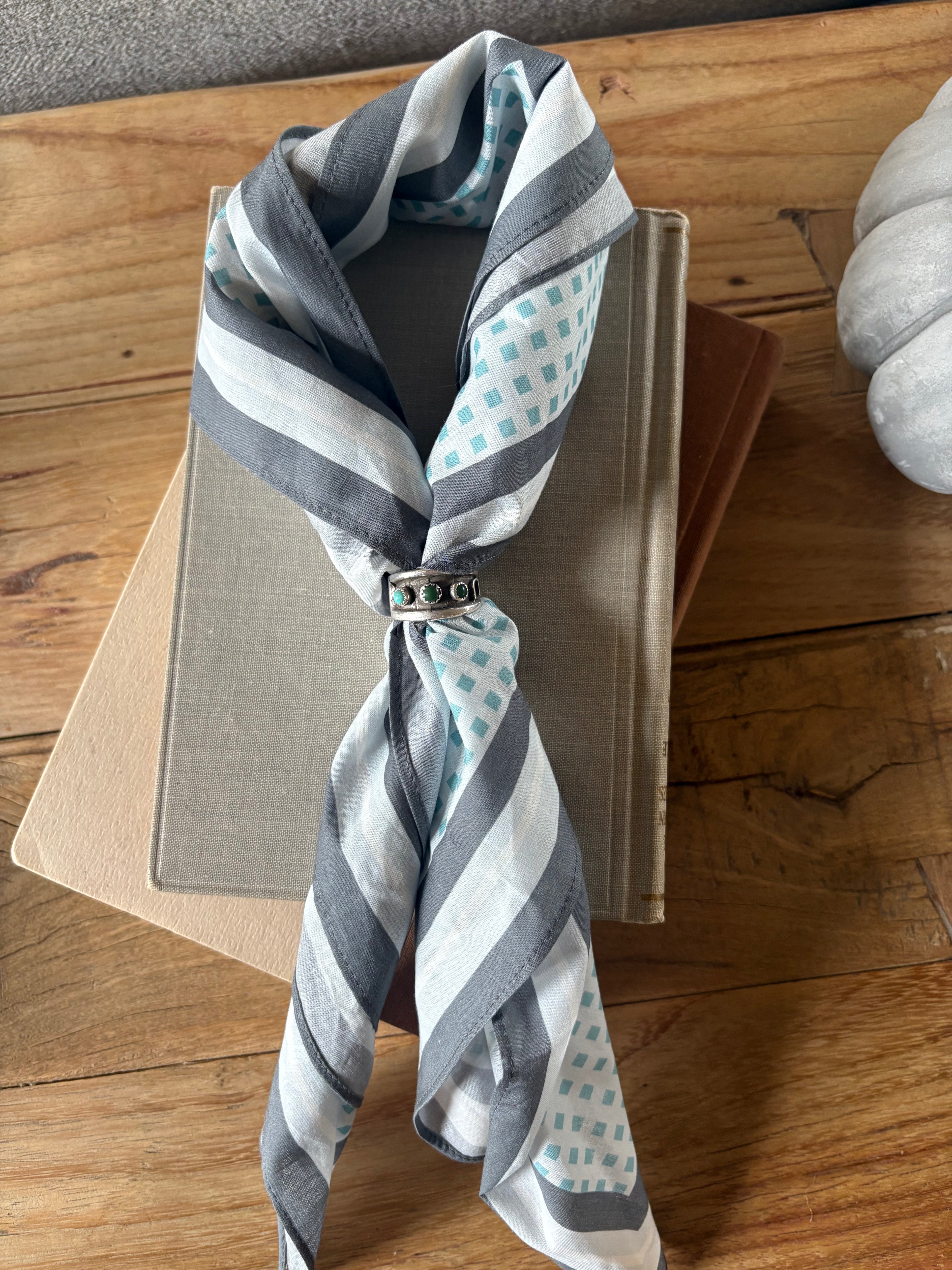 A striped silk scarf with gray, white, and light blue patterns, tied with a silver ring and draped over stacked books on a wooden table.