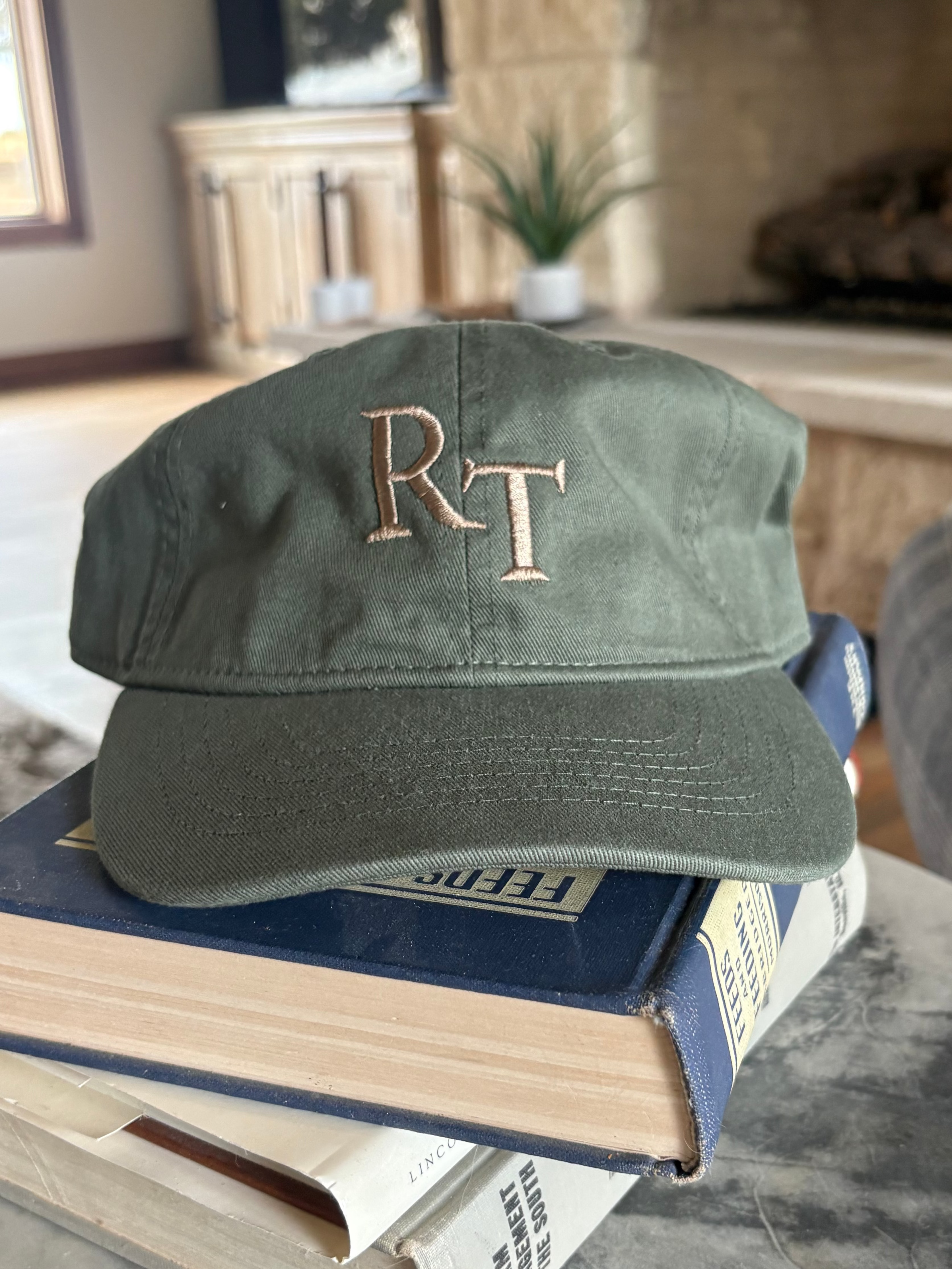 An olive green baseball cap with embroidered gold 'RT' lettering sits on top of a stack of books on a round gray marble table in a living room setting.