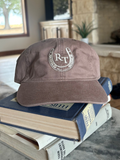 A dusty brown baseball cap with a white embroidered horseshoe and 'R&T' logo, resting on a stack of books on a stone table in a living room setting.