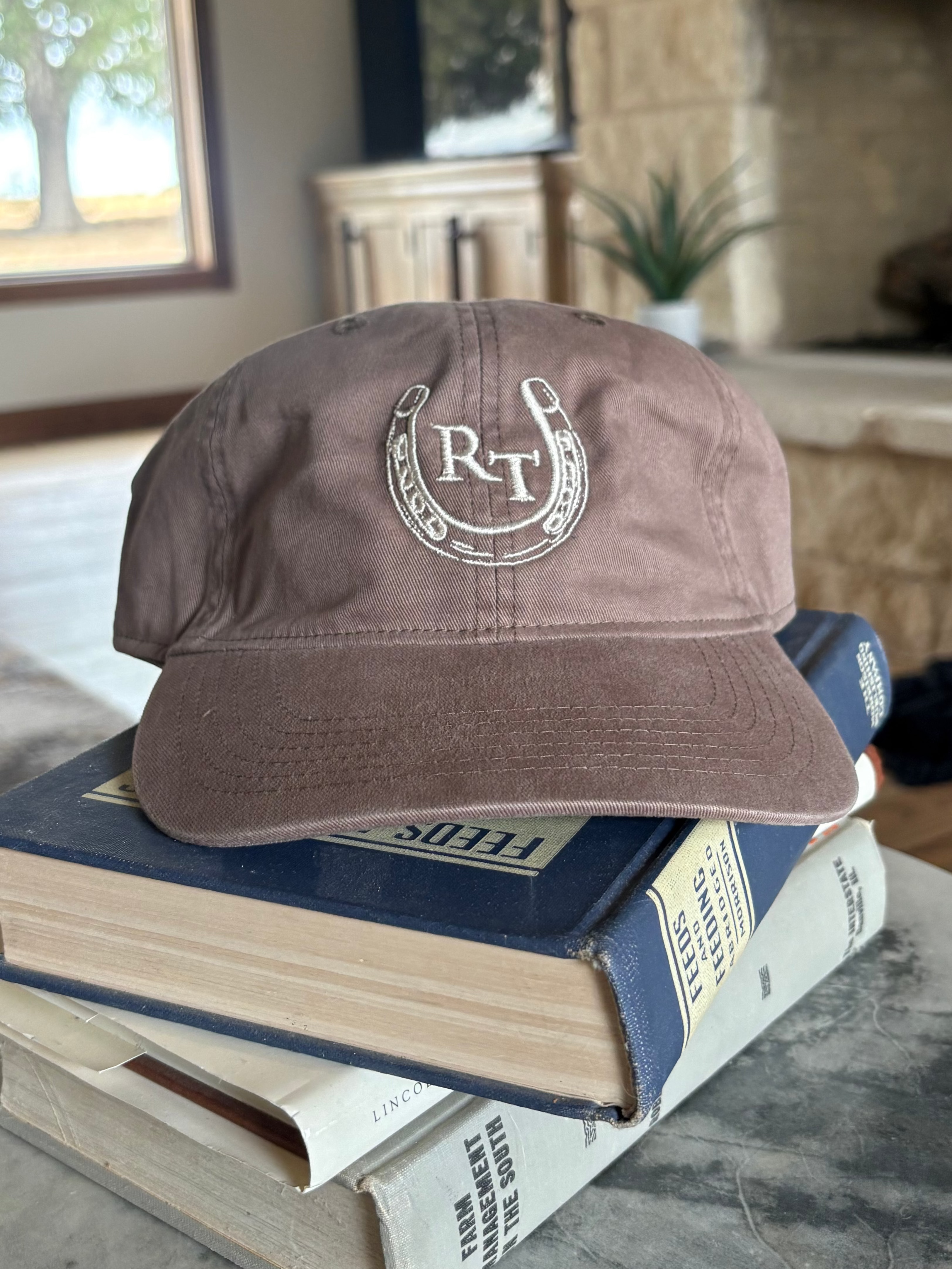A dusty brown baseball cap with a white embroidered horseshoe and 'R&T' logo, resting on a stack of books on a stone table in a living room setting.