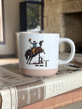 A white ceramic mug with a beige bottom half and a western-themed graphic of a cowboy on a bucking horse, resting on a stack of vintage books in front of a blurred fireplace background.