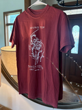 A maroon short-sleeve graphic t-shirt hanging on a white hanger in front of a large round mirror. The shirt features a white line-art graphic of a cowboy riding a bucking bronco, with the text 'ROUND TOP' above and 'BANCH GO!' below.
