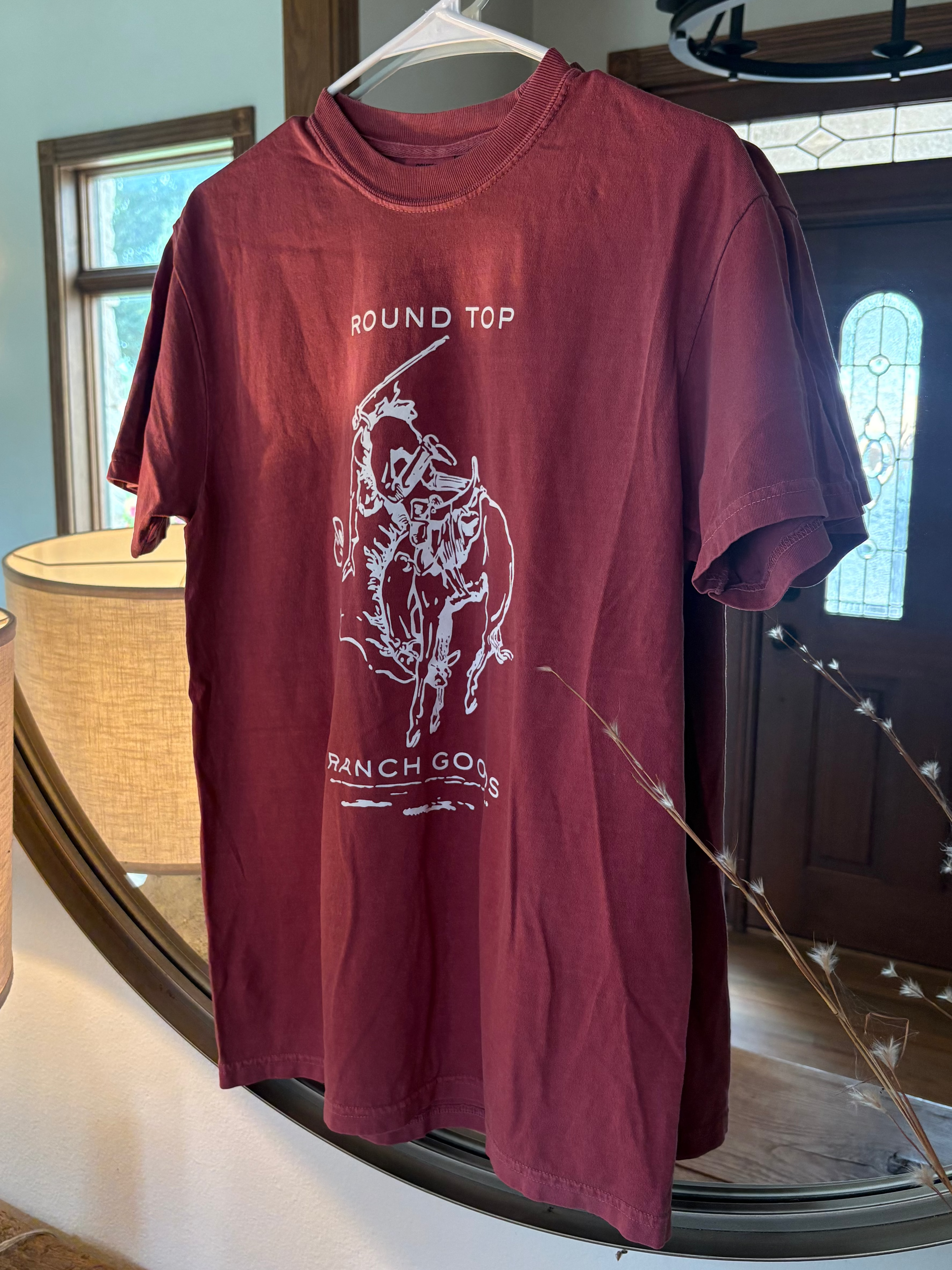 A maroon short-sleeve graphic t-shirt hanging on a white hanger in front of a large round mirror. The shirt features a white line-art graphic of a cowboy riding a bucking bronco, with the text 'ROUND TOP' above and 'BANCH GO!' below.