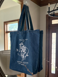A blue canvas tote bag with a white graphic print of a cowboy on a horse and the text 'ROUND TOP RANCH GOODS' hanging in front of a large round mirror inside a home entryway.