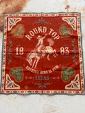 A square red bandana with a western theme, featuring a cowboy riding a bucking horse, the words 'ROUND TOP' and 'NOTHING HERE IS NEW TEXAS', and decorative heart and floral motifs in cream, green, and blue, laid flat on a textured off-white surface.