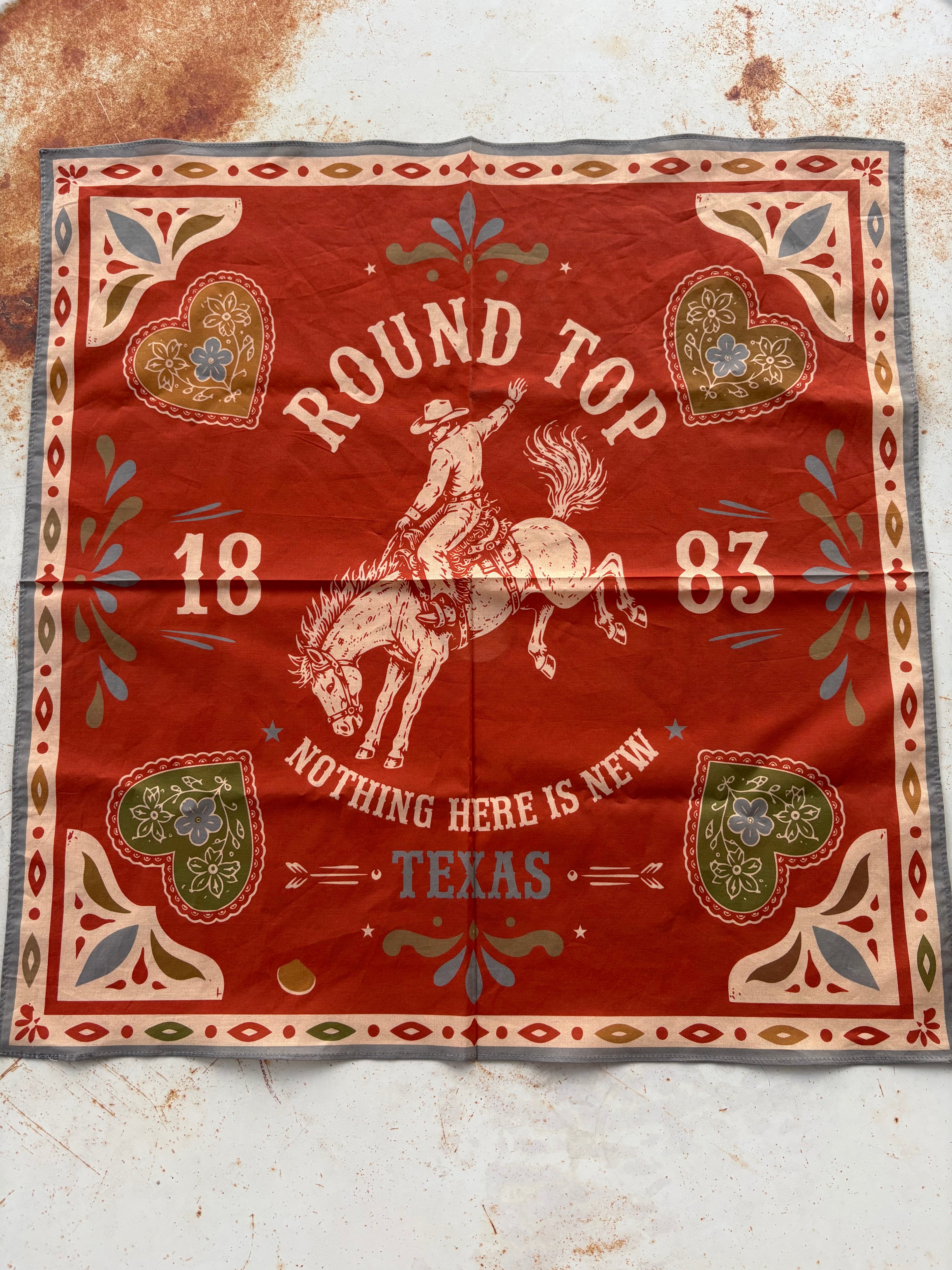 A square red bandana with a western theme, featuring a cowboy riding a bucking horse, the words 'ROUND TOP' and 'NOTHING HERE IS NEW TEXAS', and decorative heart and floral motifs in cream, green, and blue, laid flat on a textured off-white surface.